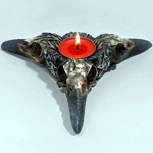 Puede incluir: Un portavelas decorativo con forma de calavera de cuervo con una vela roja encendida. El soporte está detallado con texto grabado y detalles verdes. La calavera de cuervo es de color gris oscuro y negro.