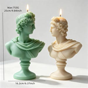 May include: Two bust candles, one sage green and one cream, shaped like classical figures with curly hair and draped fabric. Each candle has a lit wick. Text indicates a wax weight of 753g, a height of 25cm (9.84 inches), and a width of 16.2cm (6.37 inches).