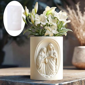 Könnte beinhalten: Eine cremefarbene, zylindrische Vase mit einem erhabenen Relief religiöser Figuren in einem ovalen Rahmen. Die Vase enthält weiße Blumen mit grünen Blättern. Ein eingesetztes Bild zeigt ein weißes zylindrisches Objekt.