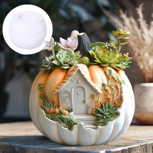 Puede incluir: Una maceta decorativa con forma de calabaza con una pequeña fachada de casa y plantas suculentas. La maceta es naranja y blanca, con un diseño detallado. La casa tiene una puerta y ventanas. La maceta está llena de varias suculentas verdes y rosas.