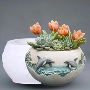 May include: A ceramic planter with a dolphin design, filled with orange tulips and succulents. The planter is off-white with a textured finish, featuring two dolphins leaping from blue waves. A white mold is next to the planter.