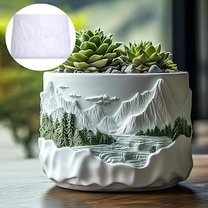 Op de afbeelding: Witte keramische plantenpot met een gedetailleerd berg- en bosreliëf. De pot bevat groene vetplanten en kleine grijze stenen. Het reliëf toont bergen, bomen, wolken en een waterval. Een decoratief item voor kamerplanten.