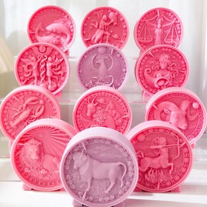Può includere: Dodici segni zodiacali rotondi in ceramica rosa e viola con disegni in rilievo. I segni sono disposti in cerchio su una superficie bianca.