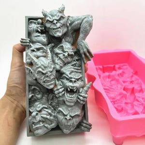 May include: A gray rectangular sculpture featuring multiple grotesque gargoyle faces emerging from a frame. The sculpture is highly detailed with sharp features and a textured surface. A pink silicone mold is visible in the background.