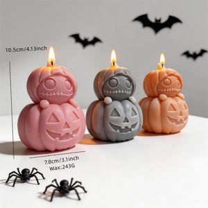 Puede incluir: Tres velas de calabaza apiladas en rosa, gris y naranja, cada una con una mecha encendida. La calabaza superior tiene una boca cosida y la inferior una cara de jack-o'-lantern tallada. Dimensiones 10,5 cm de alto, 7,8 cm de ancho, 243g.