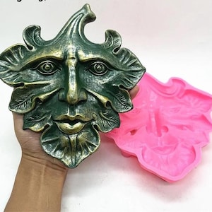 Op de afbeelding: Een groen en goudkleurige Green Man gezichtssculptuur met bladdetails. Een roze siliconen mal van hetzelfde gezicht staat ernaast. De sculptuur wordt in een hand gehouden.
