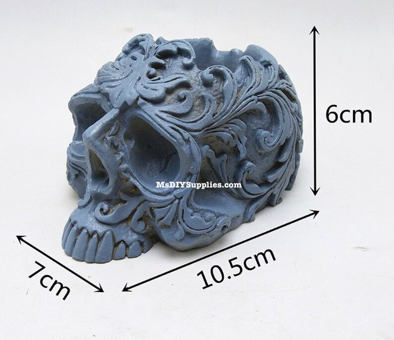 May include: A gray skull-shaped decorative container with ornate floral carvings. The container measures 4.1 inches long, 2.8 inches wide, and 2.4 inches high. The container has a hollowed-out top.