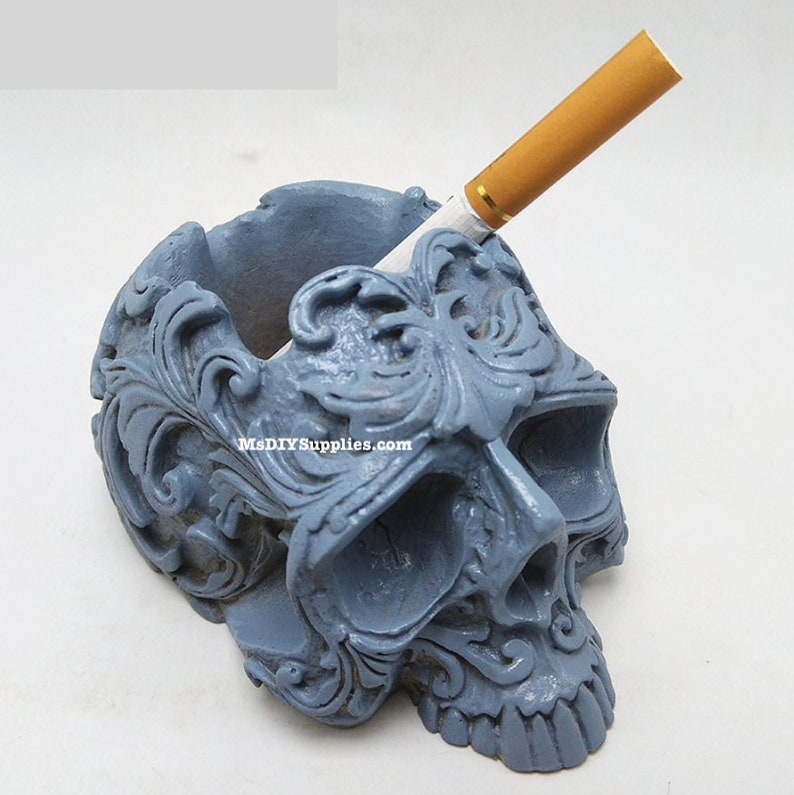 May include: A gray skull-shaped ashtray with ornate floral detailing. A lit cigarette rests in the ashtray. The text "MsDIYSUPPLIES.com" is visible on the ashtray.
