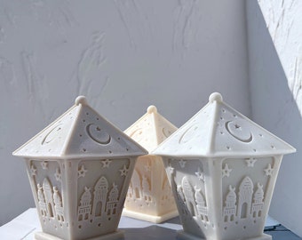 May include: Three off-white, lantern-shaped decorations. Each lantern has a pyramidal top with a crescent moon and star design. The sides feature cut-out patterns of buildings and stars. The lanterns are arranged on a white surface.