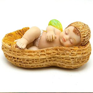 Puede incluir: Una pequeña figura de bebé hada durmiendo en una cáscara de cacahuete. El hada tiene un ala amarilla y verde y lleva un pañal blanco. La cáscara de cacahuete es marrón y tiene una superficie texturizada.