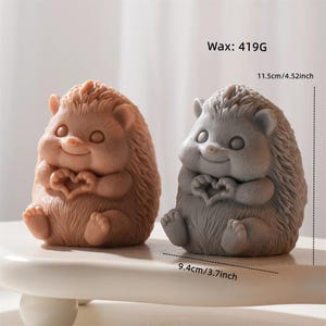 May include: Two hedgehog-shaped wax figures, one in a light brown hue and the other in gray, are displayed. Each figure is holding a heart shape with its paws. The figures are approximately 3.7 inches wide and 4.52 inches tall.