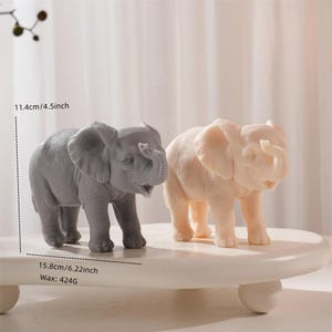 Puede incluir: Dos figuras de elefante, una gris y otra blanquecina, se exhiben sobre una plataforma blanca y redondeada. El elefante gris mide 11,4 cm de alto y 15,8 cm de largo. El elefante blanquecino es de tamaño similar. El peso de la cera es de 424g.