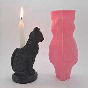 May include: A black cat candle holder with a white candle. The candle holder is made of a black resin and has a detailed cat design. The candle is lit and the flame is visible. The candle holder is sitting on a black base. The image also shows a pink silicone mold for making candles.