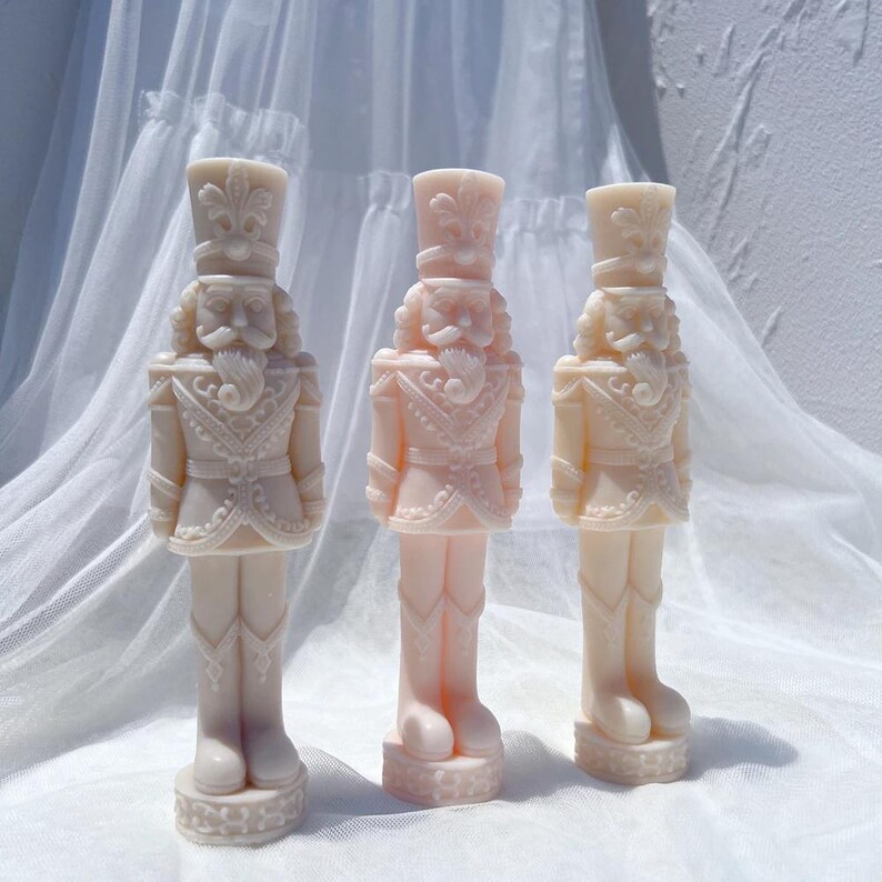 May include: Three white porcelain nutcracker figurines with intricate details. Each figurine is wearing a traditional nutcracker uniform with a hat and a beard. The figurines are standing on a white surface.