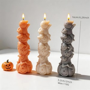 Puede incluir: Tres velas de pilar altas en naranja, crema y gris, cada una con una pila de tres figuras de fantasmas talladas sobre una base de calabaza. Una pequeña vela de calabaza naranja se encuentra cerca. Cada vela mide 16 cm de altura.