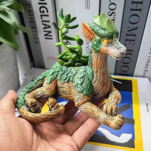 May include: A ceramic planter shaped like a mythical creature with green and brown details. The planter has a small opening on the back for planting. The planter is holding a small succulent plant.