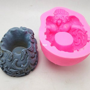 May include: A gray, ornate candle holder next to a pink silicone mold. The candle holder has a detailed, swirling design. The mold is in the shape of the candle holder, with the word "Text" visible on the mold.