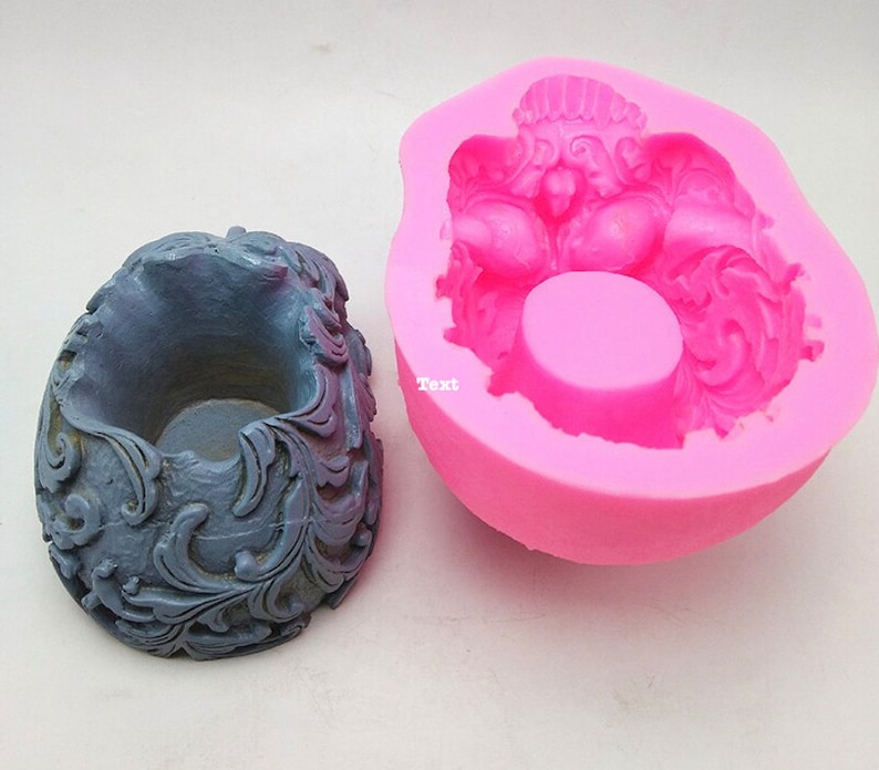 May include: A gray, ornate candle holder next to a pink silicone mold. The candle holder has a detailed, swirling design. The mold is in the shape of the candle holder, with the word "Text" visible on the mold.