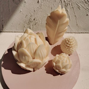 Può includere: Quattro candele bianche a forma di fiore su una superficie rosa. Le candele sono a forma di fiore, piuma, pigna e piccolo fiore.