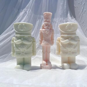 May include: Three decorative nutcracker figurines, one in pink, one in white, and one in green. The figurines are all standing upright and have a traditional nutcracker design.
