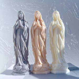 May include: Three small statues of the Virgin Mary, each in a different color: gray, beige, and white. The statues are all in a praying pose with their hands clasped together.