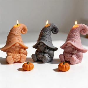 May include: Three gnome-shaped candles in orange, gray, and pink, each wearing a tall, curved hat decorated with bats. Each gnome holds a small jack-o'-lantern and a cat, with a lit wick at the top. Two small orange pumpkins sit in the foreground.