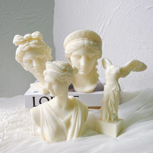 May include: A collection of off-white classical sculptures, including busts and a winged figure. The sculptures are arranged on a white surface, showcasing detailed facial features and draped garments. The pieces evoke ancient Greek and Roman art.