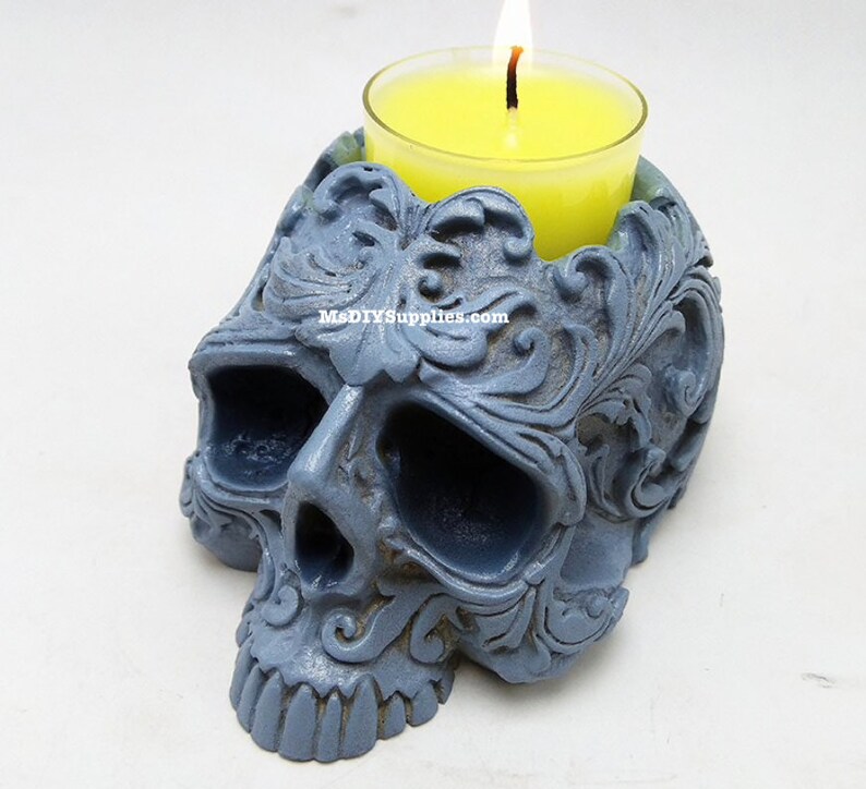 May include: A gray skull-shaped candle holder with intricate floral carvings. A yellow candle burns inside a glass votive. The candle holder has a matte finish and the text "MsDIYSupplies.com" is visible.