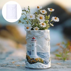 May include: A white ceramic vase with a lighthouse design, holding white daisy flowers with yellow centers. The vase features a detailed relief of a lighthouse, ocean waves, and coastal plants. The vase is approximately 8 inches tall.
