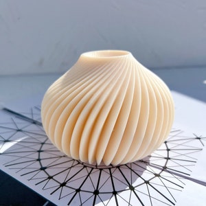 以下が含まれることがあります： 渦巻き状の肋骨模様が施された白い陶器の花瓶。花瓶は丸く、上部に広い開口部があります。
