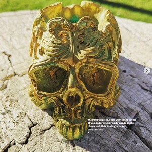 May include: A gold-colored skull-shaped decorative item with intricate, ornate details. The skull has large eye sockets, a prominent nose, and a detailed jawline. The surface has a textured appearance with hints of green. The item is sitting on a weathered wooden surface.