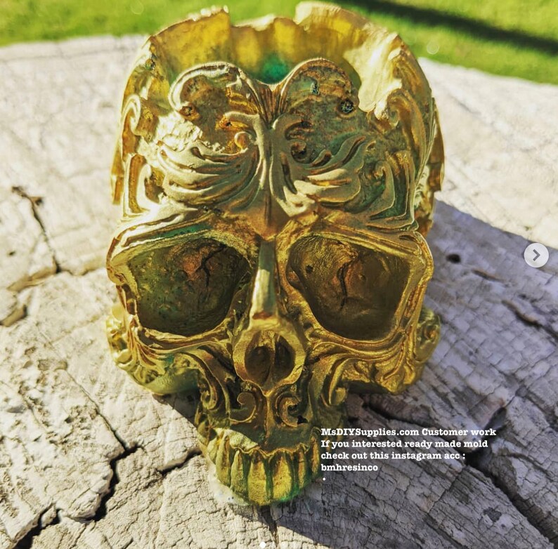 May include: A gold-colored skull-shaped decorative item with intricate, ornate details. The skull has large eye sockets, a prominent nose, and a detailed jawline. The surface has a textured appearance with hints of green. The item is sitting on a weathered wooden surface.