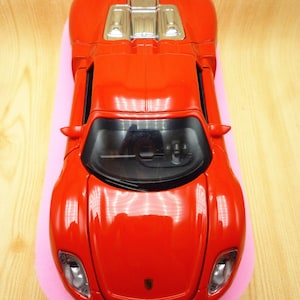 May include: A red toy car with a silver engine detail. The car is on a pink surface.