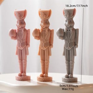Könnte beinhalten: Drei hasenförmige Figuren in Rosa, Pfirsich und Grau, die jeweils ein Ei halten. Die Figuren sind 18,2 cm hoch und 5 cm breit, mit einem Sockel. Sie sind in Uniformen und Hüten im Militärstil gekleidet.