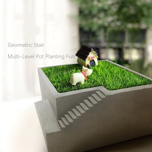 Puede incluir: Una jardinera de hormigón gris con un diseño de escalera geométrica. La jardinera contiene césped verde artificial, una casa en miniatura y una pequeña figura de perro. El texto en la imagen dice "Geometric Stair" y "Multi-Level Pot Planting Fun."