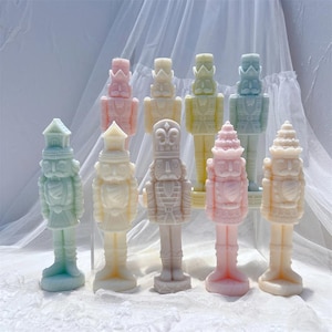 Puede incluir: Una colección de figuras de cascanueces de colores pastel. Las figuras son en tonos de rosa, amarillo, verde y blanco. Cada cascanueces tiene un uniforme y un sombrero detallados. El fondo es una tela blanca suave.