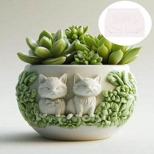 Può includere: Vaso in ceramica bianca con un design floreale dettagliato e due gattini bianchi scolpiti. Il vaso è pieno di piante grasse verdi.