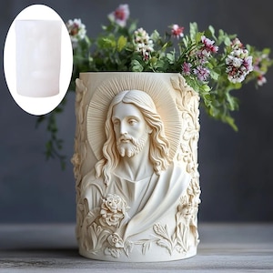 Könnte beinhalten: Eine cremefarbene, zylindrische Vase mit einem detaillierten Relief von Jesus Christus. Die Vase hat einen Heiligenschein und florale Akzente und ist mit grünen und lila Kunstblumen gefüllt. Die Vase besteht aus einem haltbaren Material.