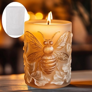 May include: A lit, cylindrical candle with a detailed bee design. The candle is a warm, amber color, with a sculpted bee and leaf pattern. The flame is bright, and the candle appears to be made of wax. A white, cylindrical container is in the upper left corner.