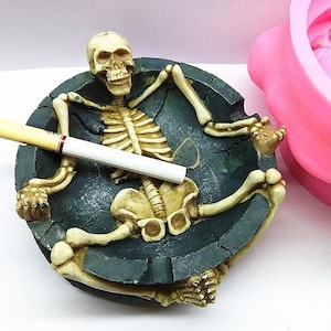 May include: A novelty ashtray featuring a skeleton figure reclining in a dark green, cracked-effect dish. A lit cigarette rests across the skeleton's chest. The ashtray is made of a beige and green material.