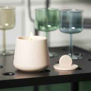 May include: A white ceramic candle with a lit wick sits on a black surface with three clear glass cups. The candle has a lid with a small knob.