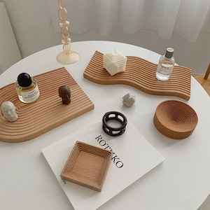 May include: A set of wooden trays and bowls with a minimalist design. The trays have a wavy shape and are made of light wood. The bowls are round and have a concentric circle pattern. There are also two small sculptures of human heads, a candle, and a bottle of perfume on the trays.