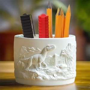 May include: A white ceramic pencil holder with a detailed relief of a dinosaur and landscape. The holder contains a black pen, a red eraser, and several yellow pencils. The design features mountains, trees, and a building.