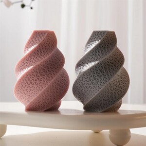 May include: Two decorative spiral-shaped vases. One is a soft pink, the other is gray. Both have a textured surface with a repeating geometric pattern. They sit on a white, rounded-edge platform.