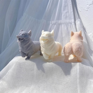 Puede incluir: Tres figuritas de perro de cerámica en diferentes colores: gris, blanco y rosa. Las figuritas están todas en la misma pose, con la cabeza inclinada hacia un lado y la lengua fuera. Están sentadas sobre una superficie de tela blanca.