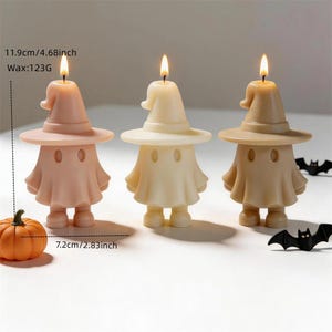 May include: Three lit candles shaped like ghosts wearing witch hats in light pink, cream, and tan. Each candle is 4.68 inches tall and weighs 123 grams. A small orange pumpkin and two black bat figures are placed near the candles.