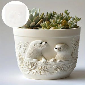 May include: Cream ceramic plant pot featuring a relief of two otters. The pot is filled with green and light green succulents. The otters are finely detailed, and the pot has a textured surface. The pot is suitable for indoor use.