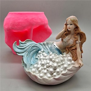 Puede incluir: Una detallada figura de sirena en una concha, con una cola azul claro y un top blanco y plateado. La sirena tiene un largo cabello rojizo y está rodeada de burbujas blancas con forma de perlas. Un molde de silicona rosa está en el fondo.