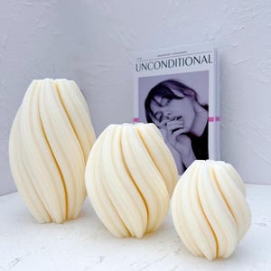 May include: Three white, decorative candles with a swirling, ribbed design. The candles are arranged in a row from largest to smallest.