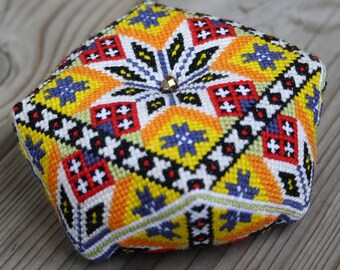 May include: A colorful, geometrically patterned, square-shaped pincushion with a gold button in the center.
